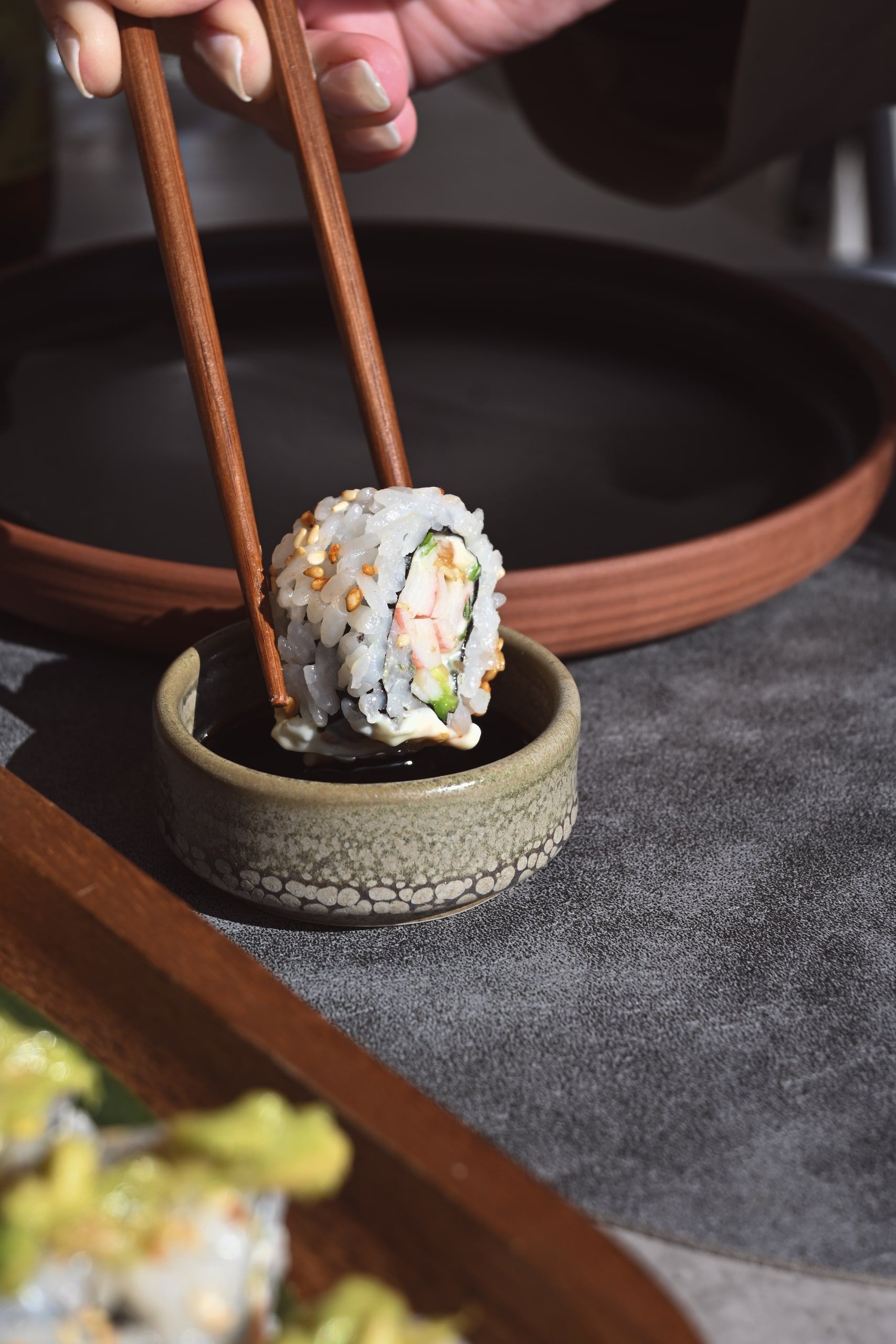 Ρολό sushi ανάμεσα σε ξύλινα chopsticks πάνω από κεραμικό πιατάκι, σε σκούρο φόντο.