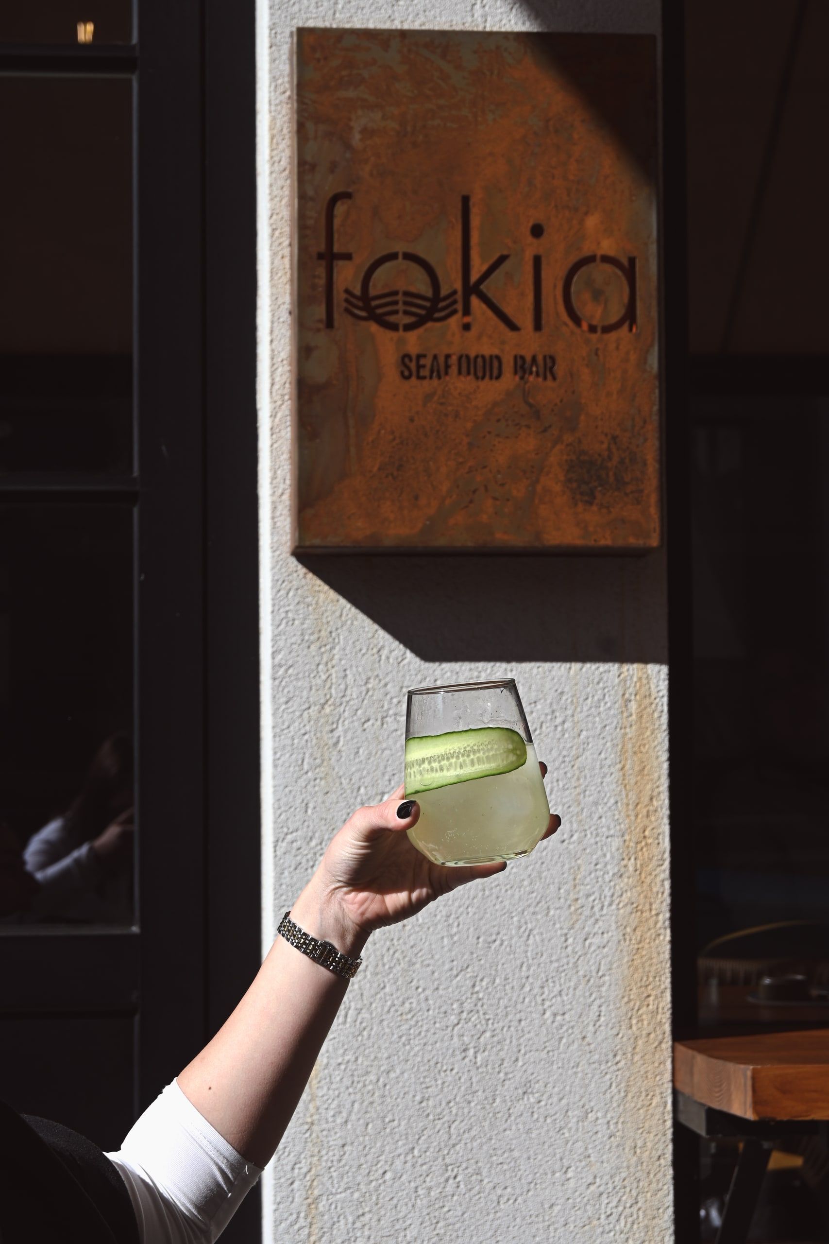 Η πινακίδα «fokia — seafood bar» από οξειδωμένο ατσάλι στην πρόσοψη του εστιατορίου, με ένα χέρι να κρατά ένα δροσιστικό κοκτέιλ.