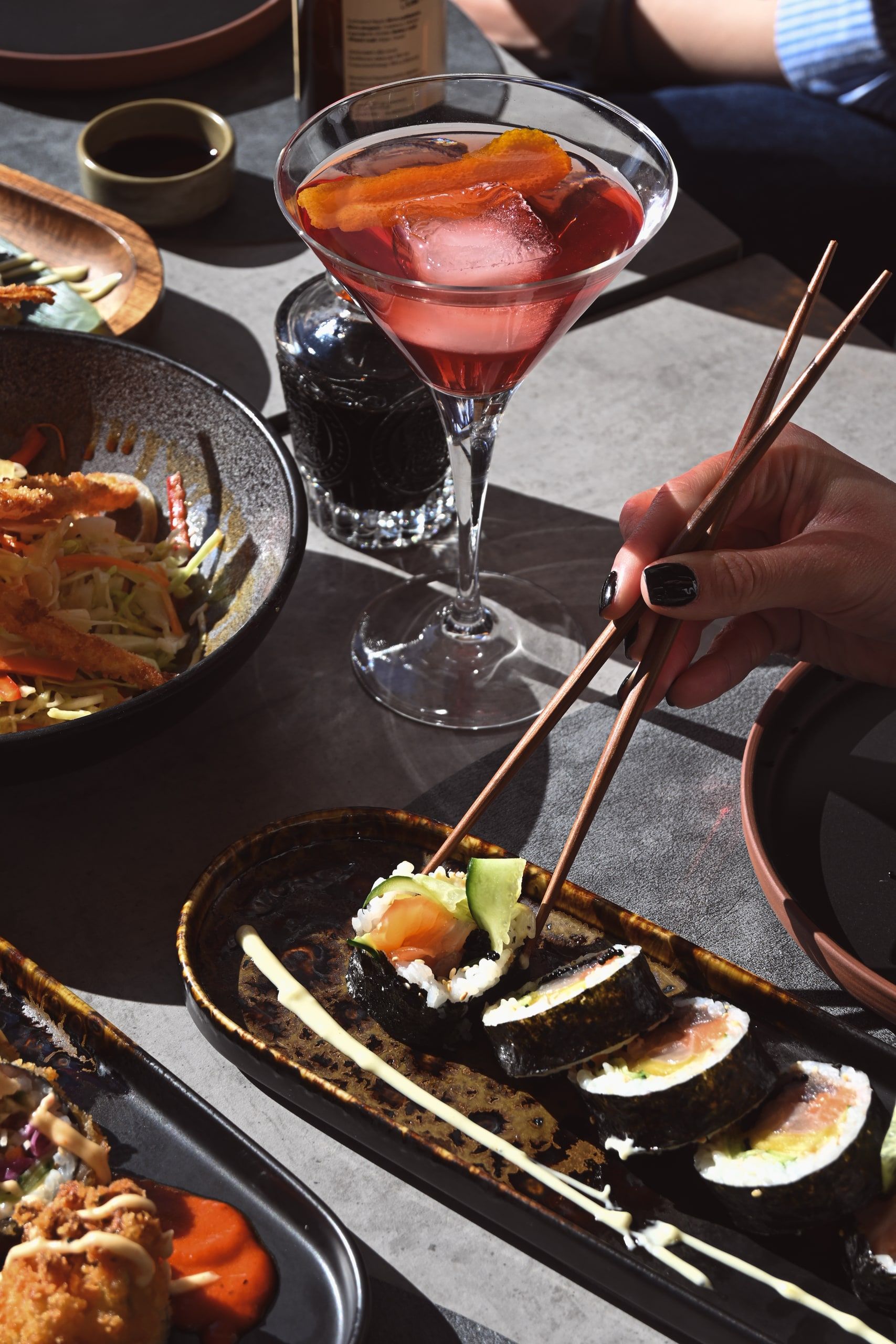 Χέρι με chopsticks απλώνεται προς ρολά maki σε σκούρα πλατέλα, δίπλα σε κοκτέιλ.
