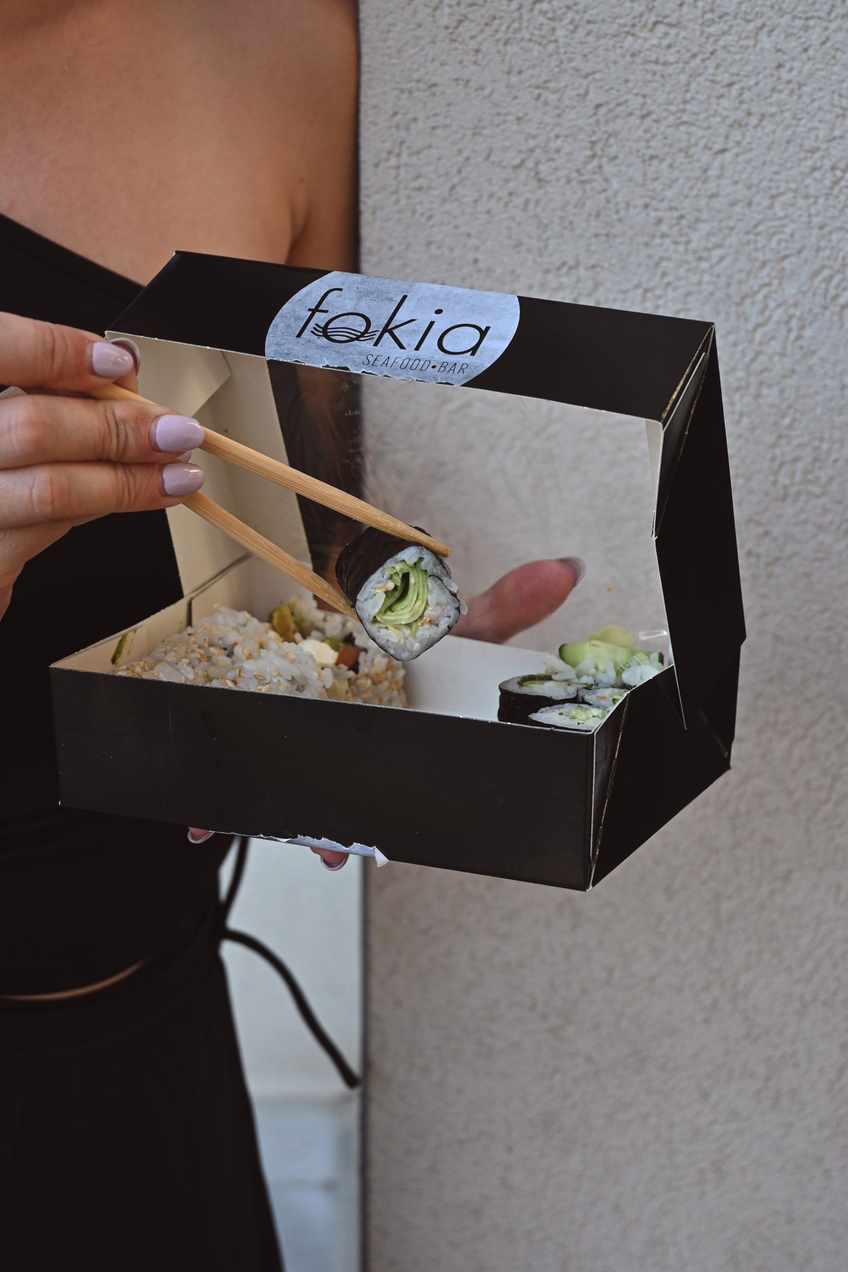 Χέρι κρατά κουτί takeaway με το λογότυπο fokia, με ένα ρολό sushi πιασμένο με chopsticks.