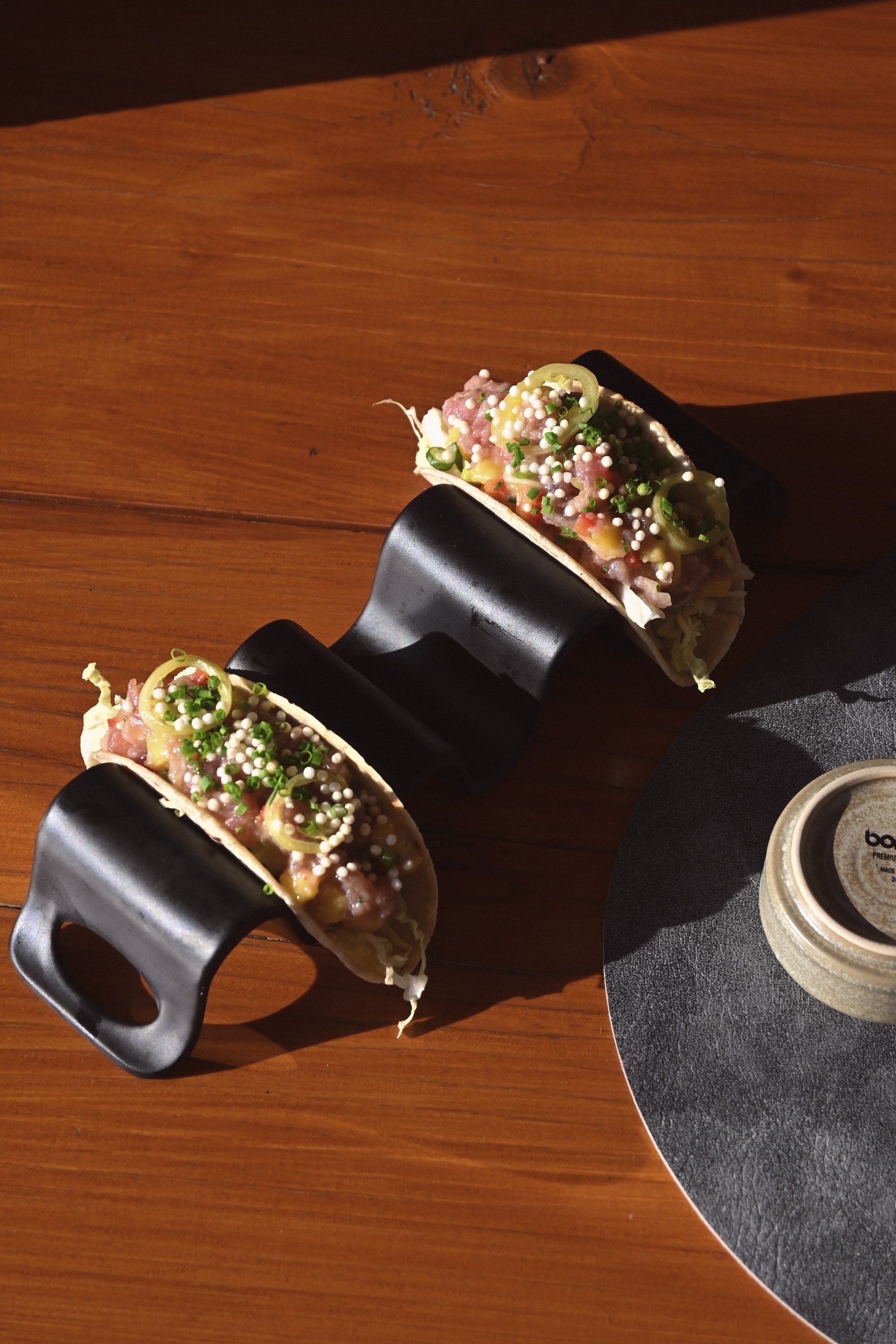 Δύο tacos σε μαύρες βάσεις σερβιρίσματος πάνω σε ξύλινο τραπέζι στο ηλιακό φως.