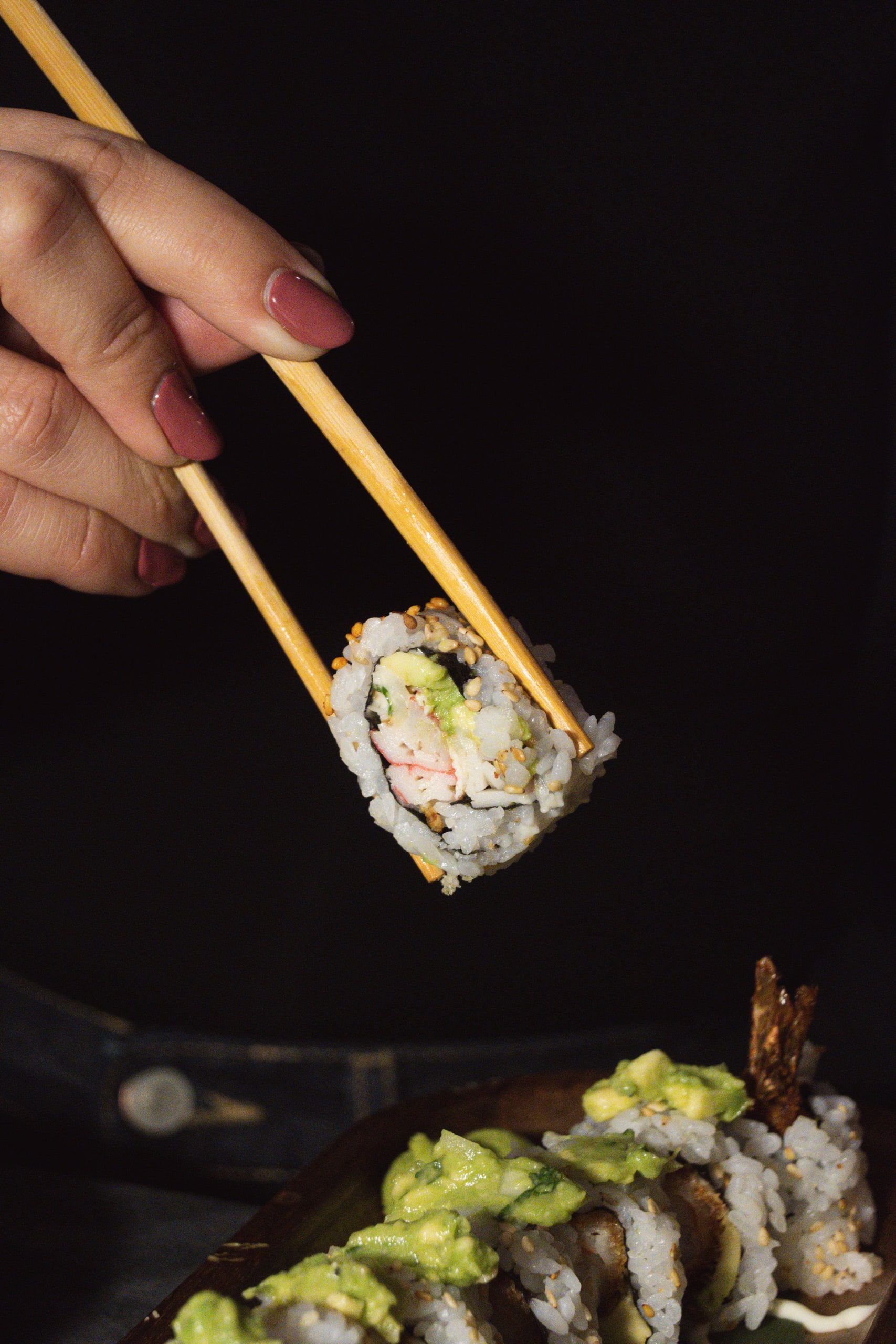 Κοντινό πλάνο ρολού California πιασμένου με chopsticks πάνω από το τραπέζι.